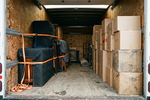 Blanket-wrapped furniture held upright by orange ratchet straps through the wall rails of a loaded moving van, boxes stacked opposite and the floor clear