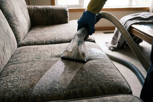 An upholstery tool drawn across a fabric sofa cushion, dirty water visible in the clear head and a clean lane behind it